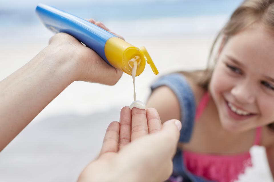 A person squeezes sunscreen out of a container in front of a smiling girl in her togs.