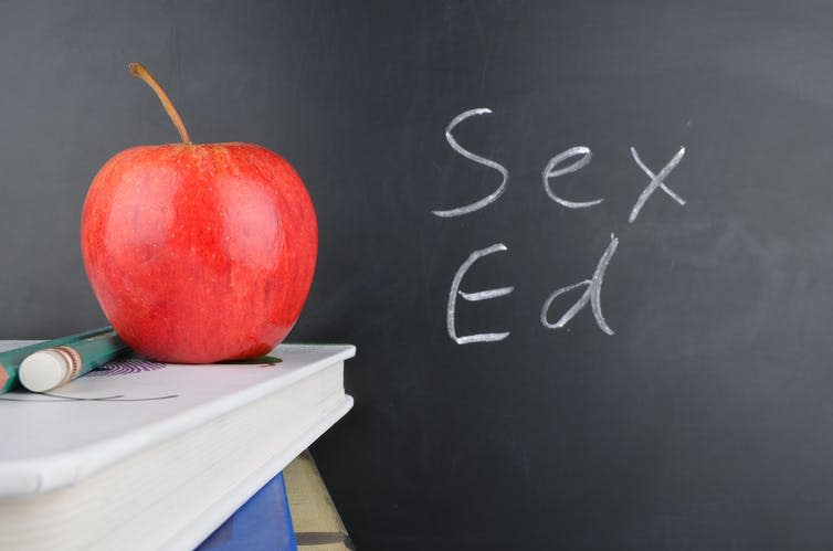 Apple and two pencils on pile of books next to blackboard that says 'Sex Ed'