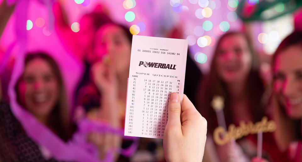 A stock image of a group of friends celebrating in the background while a woman holds up a Powerball ticket in the foreground.