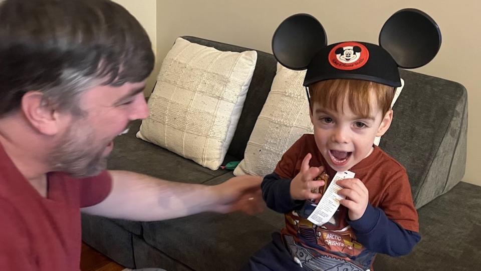 A young boy sits on a small couch with a Mickey Mouse ears hat as hid dad smiles at him.