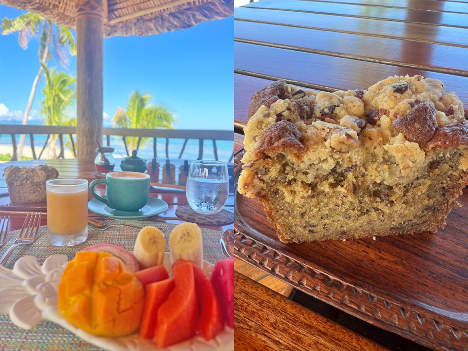 Breakfast of mango, watermelon, bananas on a plate with glass of orange juice, a blue coffee cup, a glass of water, and a slice of banana bread on a table in front of a beach; A slice of banana bread with a crumble and chocolate chips on top