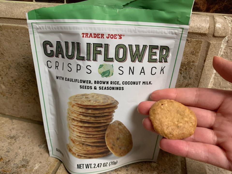 a hand holding a Trader Joe's cauliflower crisp with the original white and green bag in the background