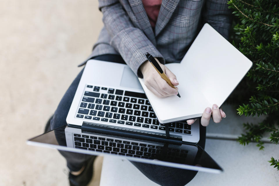 Person working on a laptop outdoors, writing in a notebook resting on the laptop's keyboard