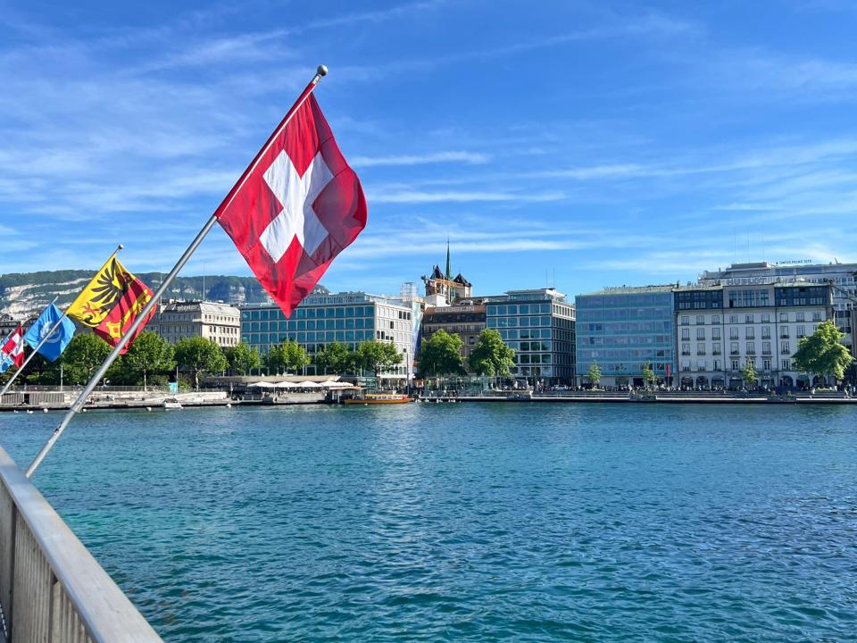 The flags of Switzerland, the Canton of Geneva, and the UN on a bridge over the Rhône with buildings in the background on a sunny day