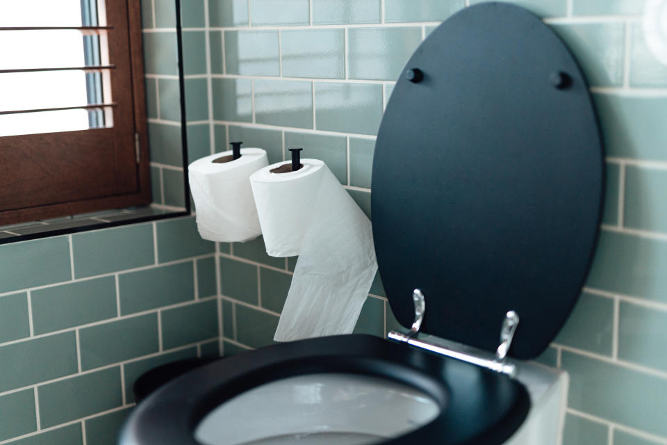 Toilette mit schwarzer Brille und Deckel vor türkisfarbenen Fliesen.