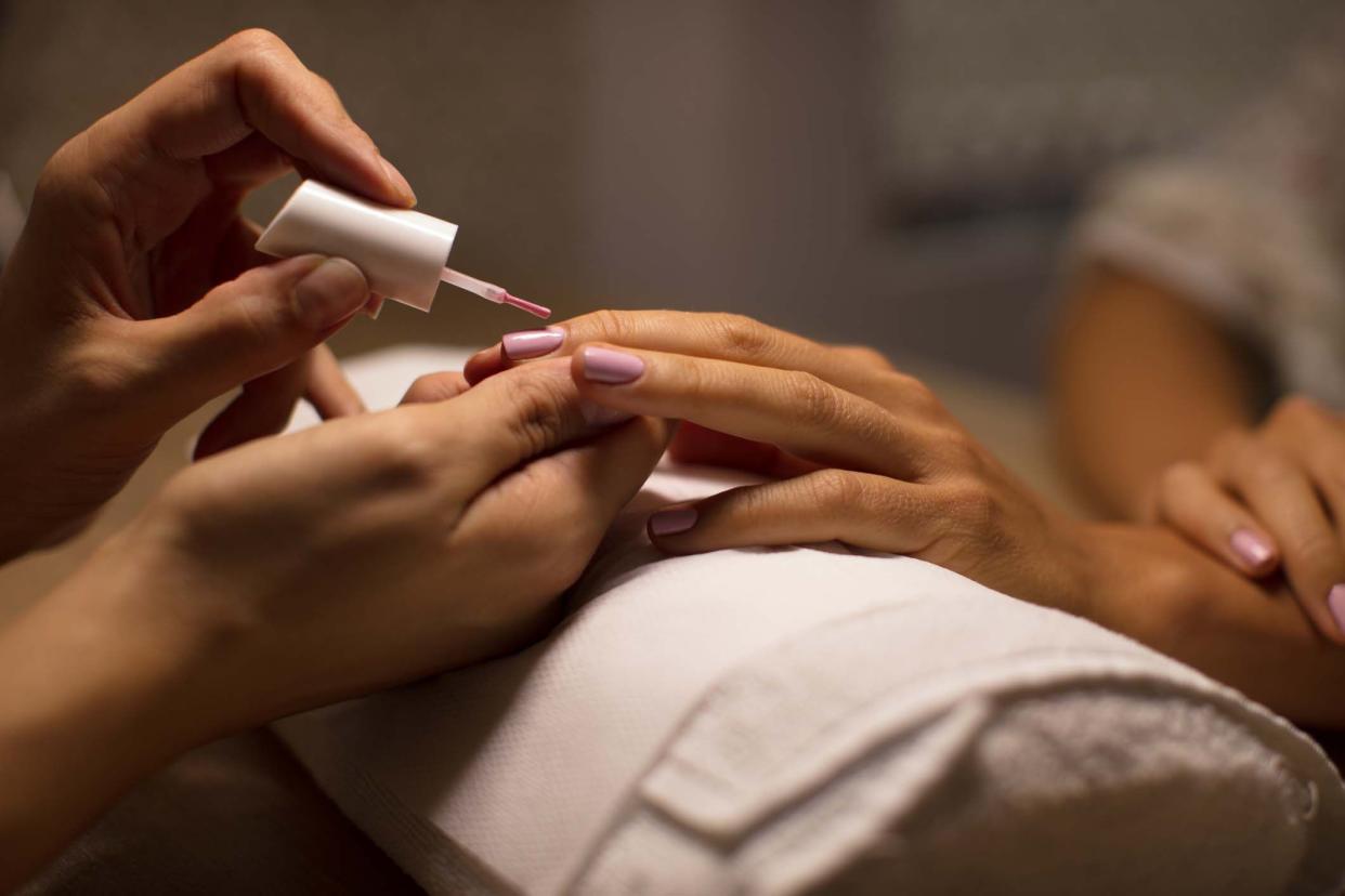 Closeup of woman's left hand getting a manicure, nails being painted a mauve pink, female nail technician's both hands, surrounded by shading