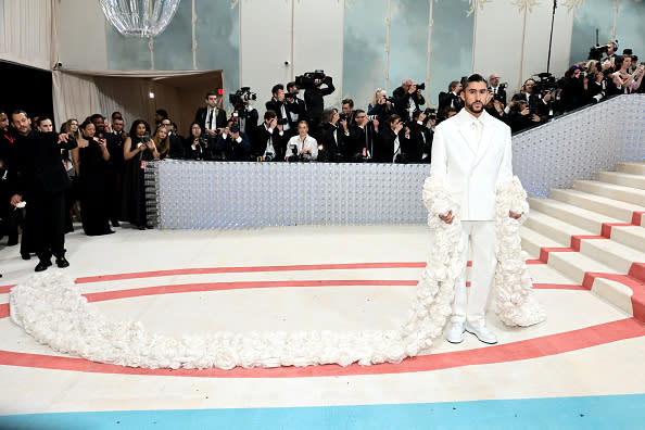 Man in ornate white outfit with fluffy accents on a staircase, photographers in background