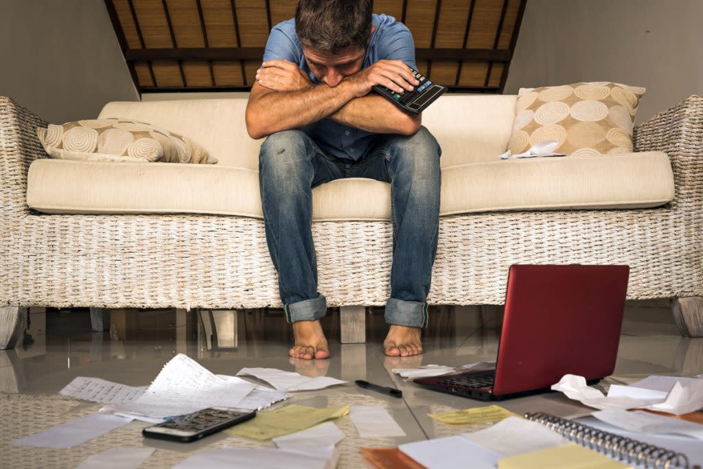 A man looks stressed with his finances spread across the floor next to a calculator and laptop. 
