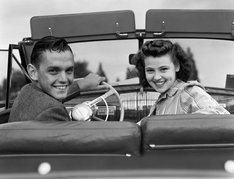A black and white image shows two teenagers sit in a 1940's convertible. They are smiling at the camera. 