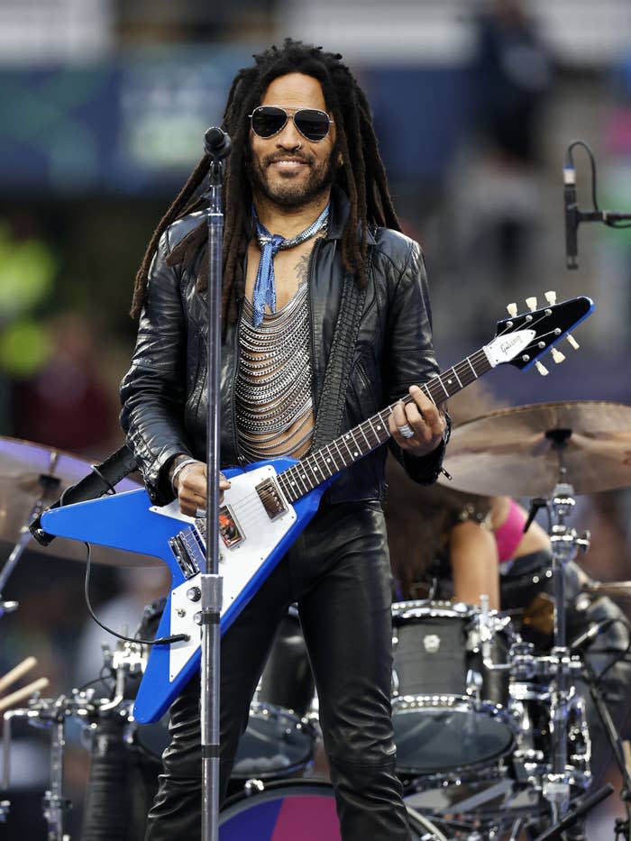 Lenny Kravitz performs on stage wearing a leather jacket, a metallic necklace, sunglasses and playing a blue and white electric guitar