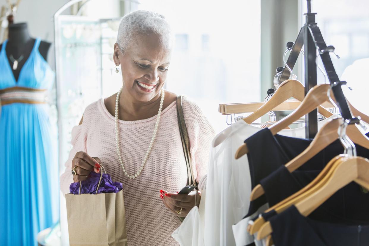 A senior African American woman shopping in a clothing store. She is standing by a rack of dresses on hangers, looking at a price tag, carrying a shopping bag.