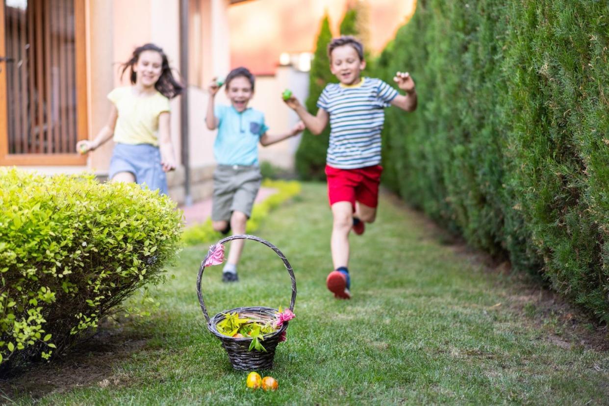 three children holding easter eggs happily racing toward basket outdoors during easter egg hunt relay