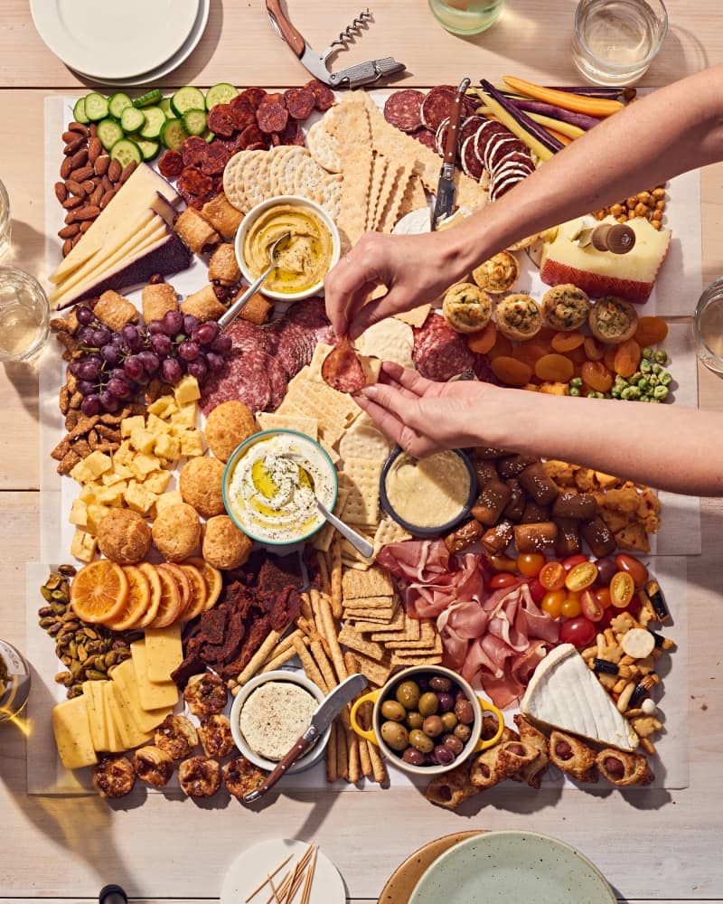 grazing table or board on a light wood table. There is meat, cheese, crackers, fruit, vegetables, dips, olives, and more. Plates and glasses, toothpicks on the table by the food. Hands are reaching in to get snacks.