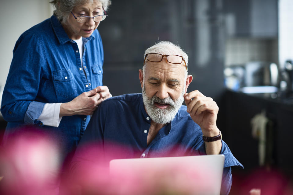 Candid portrait of senior couple at home, man with grey hair and beard working on computer, glasses resting on forehead, seniorpreneur working from home with wife