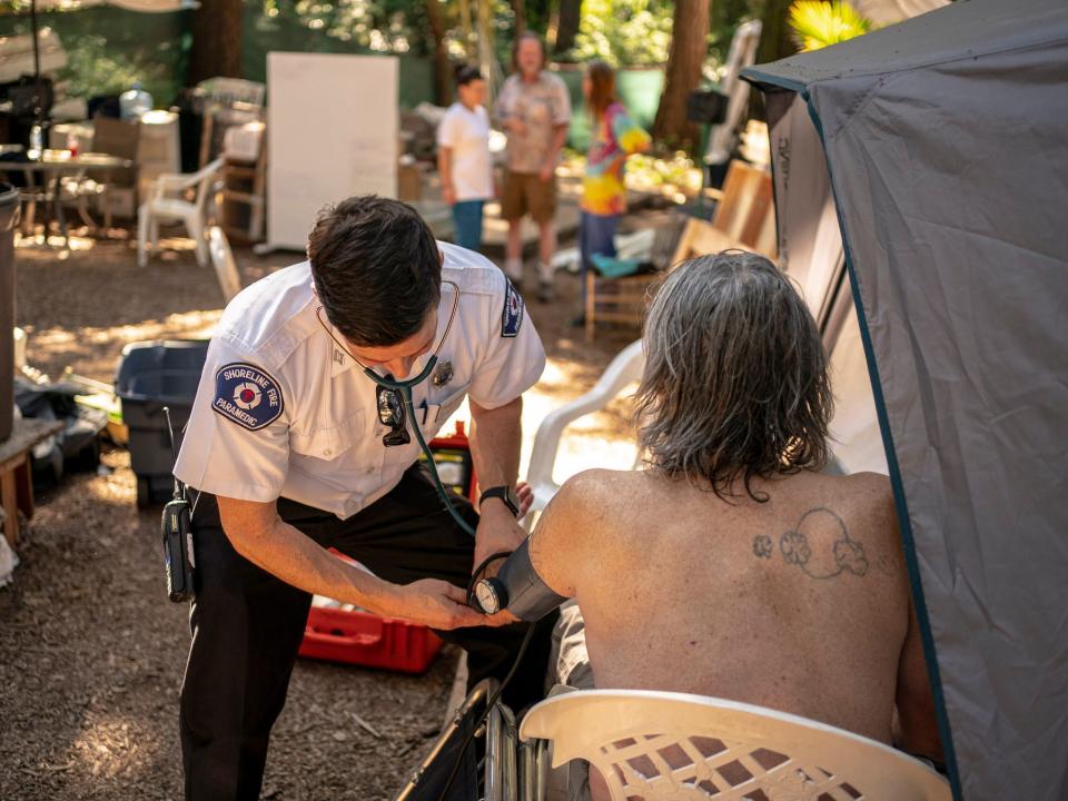 healthcare worker in t shirt uniform checks blood pressure of shirtless man in tent encampment