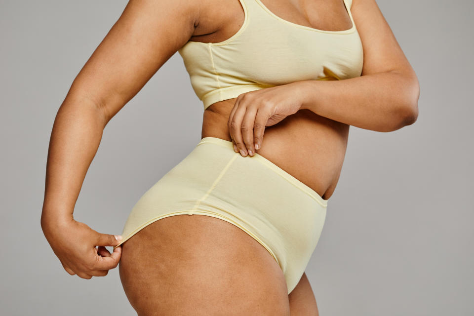 Woman in a yellow tank top and high-waisted briefs, standing with her side to the camera, pulling on her waistband