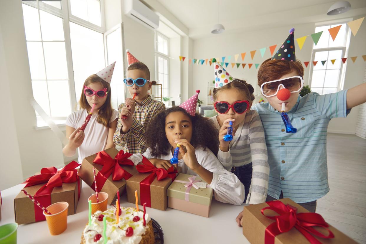 Group of happy diverse children in party hats and funny sunglasses blowing noisemakers and looking at camera while having fun during birthday celebration at home