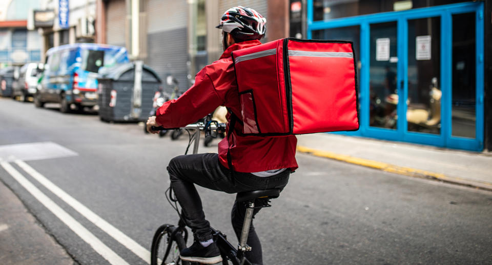 Rear view of ddelivery man, who's wearing cycling helmet and red jacket, riding bicycle on city street on his way to customer