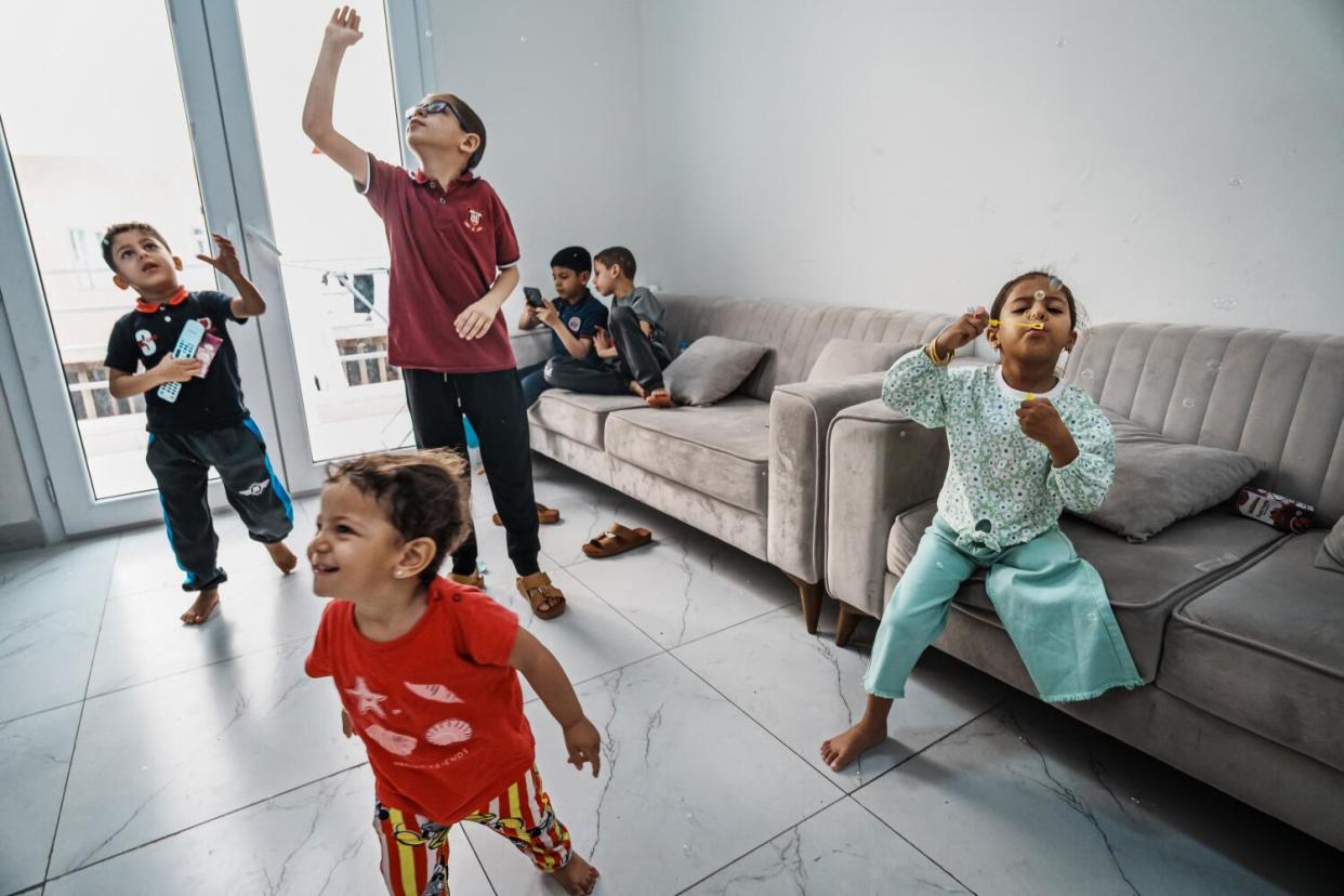 Six children blow bubbles while playing on a tile apartment floor or on couches.