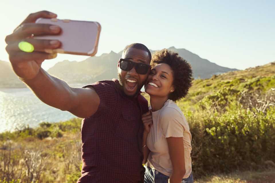 Smiling couple taking selfie in front of lake, with mountains in the background.