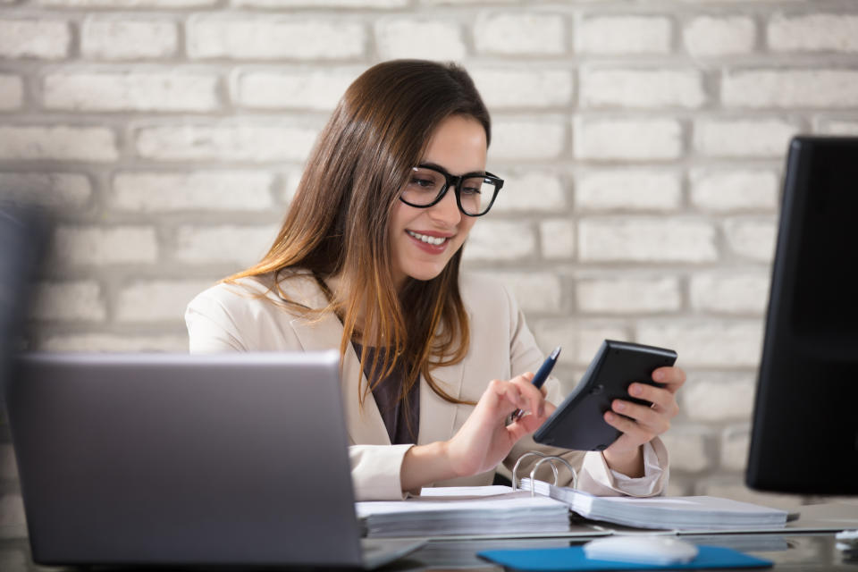 Professionally dressed woman at desk looking at calculator with binder open in front of her