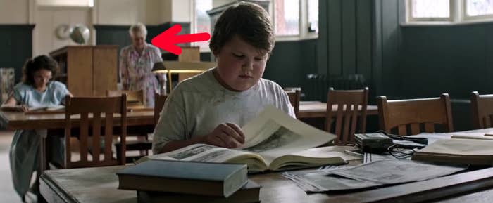 A woman wearing a sinister smirk looms behind a boy sitting at a table in the library