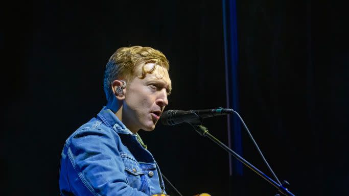 tyler childers sings into a microphone on a stand and plays acoustic guitar, he wears a jean jacket