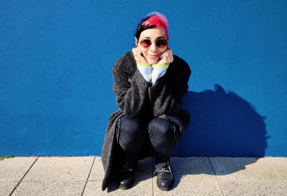 Julieta, a woman in and a coat sunglasses with pink and black hair, squatting in front of a blue wall - Courtesty of interviewee/Mialgros Costabel