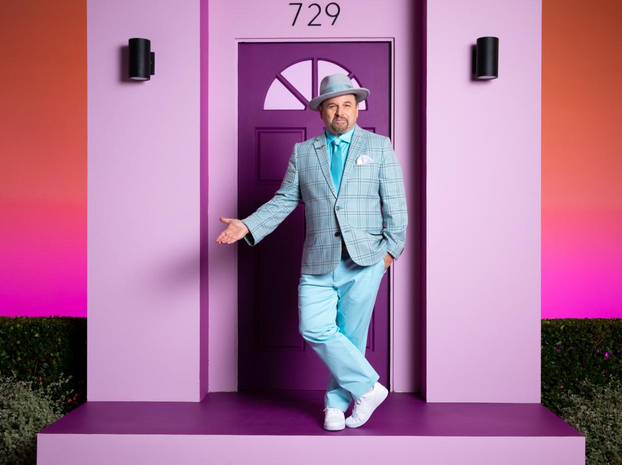 Jason Alexander standing on the porch of a purple house wearing a hat