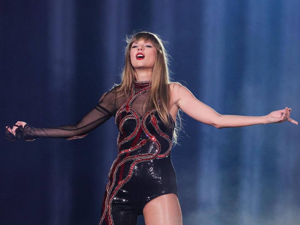 Taylor Swift extending her arms on stage while lifting her head up and wearing a black dress with pink detailing.