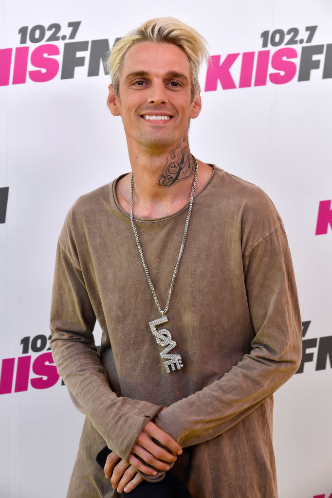 Aaron standing in front of promotional backdrop, wearing a necklace with "LOVE" pendant and casual attire