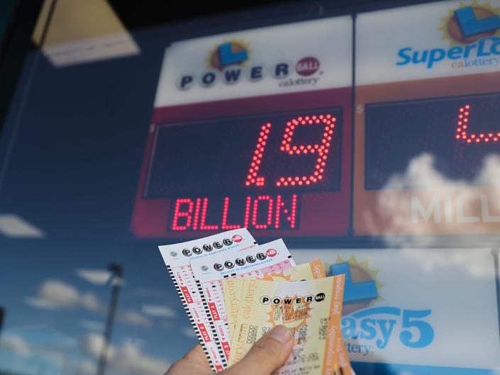 Цифровой знак рекламирует джекпот Powerball в размере 1,9 миллиарда долларов, а рука держит лотерейные билеты.