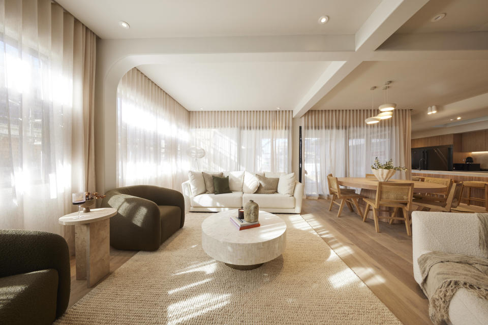 A wide shot of the living room with a round table in the middle, a couch on the back wall and sheer white curtains all around. 