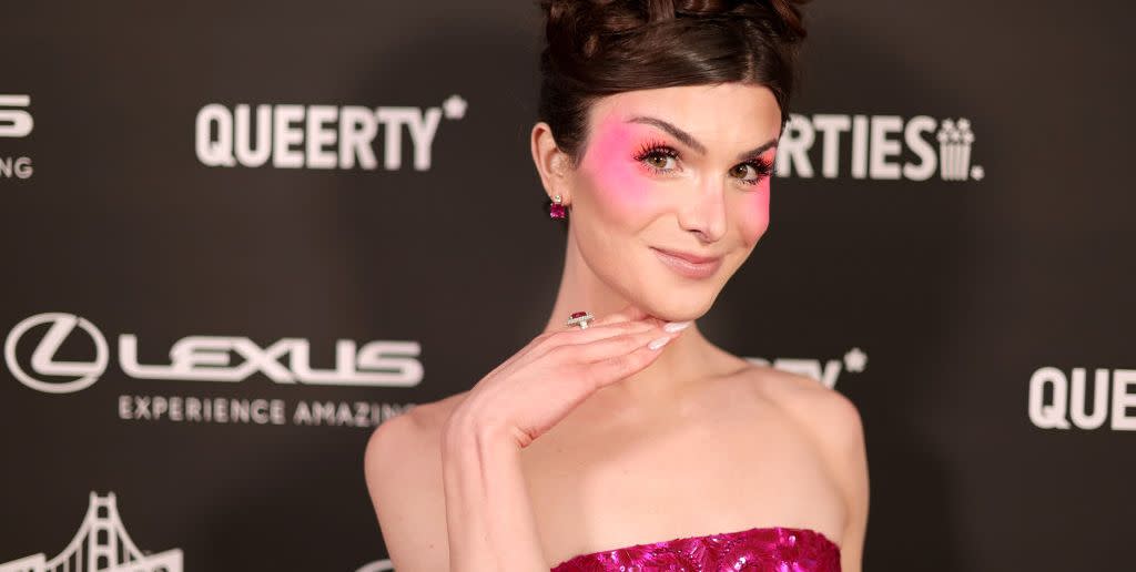 dylan mulvaney wearing a pink dress and blush makeup, putting her right hand under her chin, smiling directly at the camera