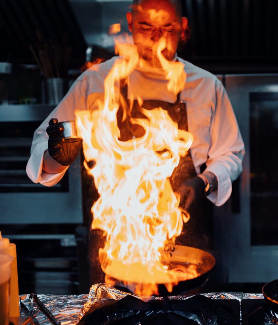 A chef in a professional kitchen uses a burner to create a large flame over a skillet. Another pan with tomatoes is visible next to him