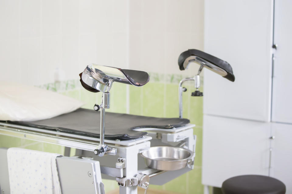 Gynecological examination table with stirrups in a medical clinic. White pillow on the table and nearby sink