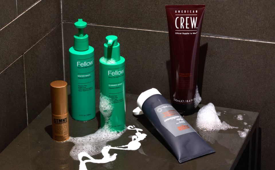 Variety of men's haircare products on a shower bench with suds and soap surrounding the products.