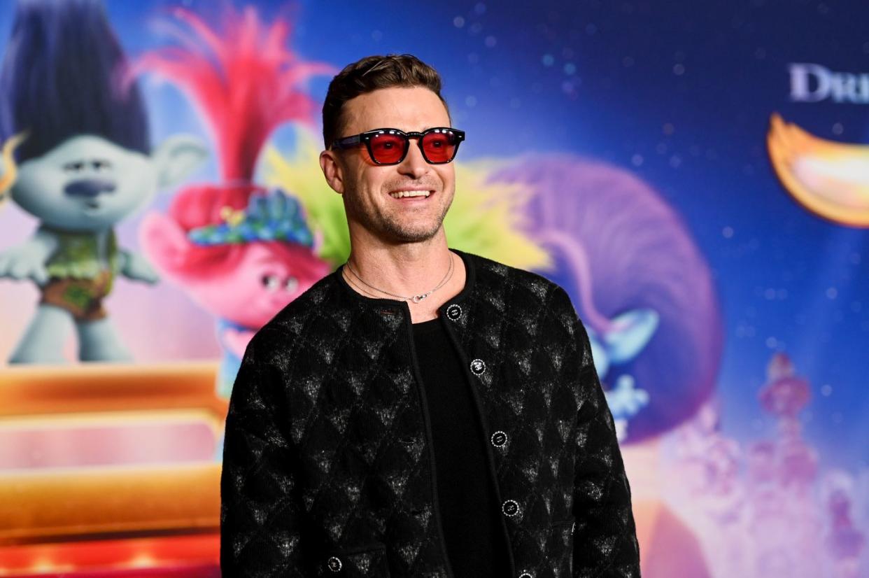justin timberlake smiles and looks right of the camera, he stands in front of a multicolor movie poster and wears a black and silver patterned jacket with a black top, red tinted glasses and a thin chain necklace