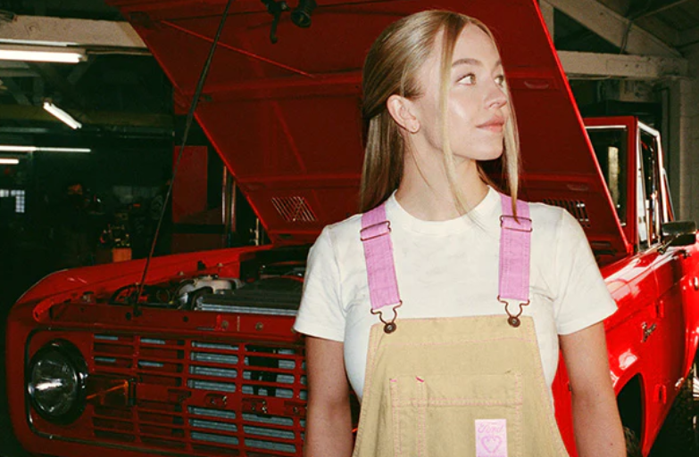 Actress Sydney Sweeney wears the bib overalls (standing in front of her 1969 Ford Bronco) that have sold out for $79.99 and are now available for pre-order at https://merchandise.ford.com/products/fordxsydneysweeney-overalls.