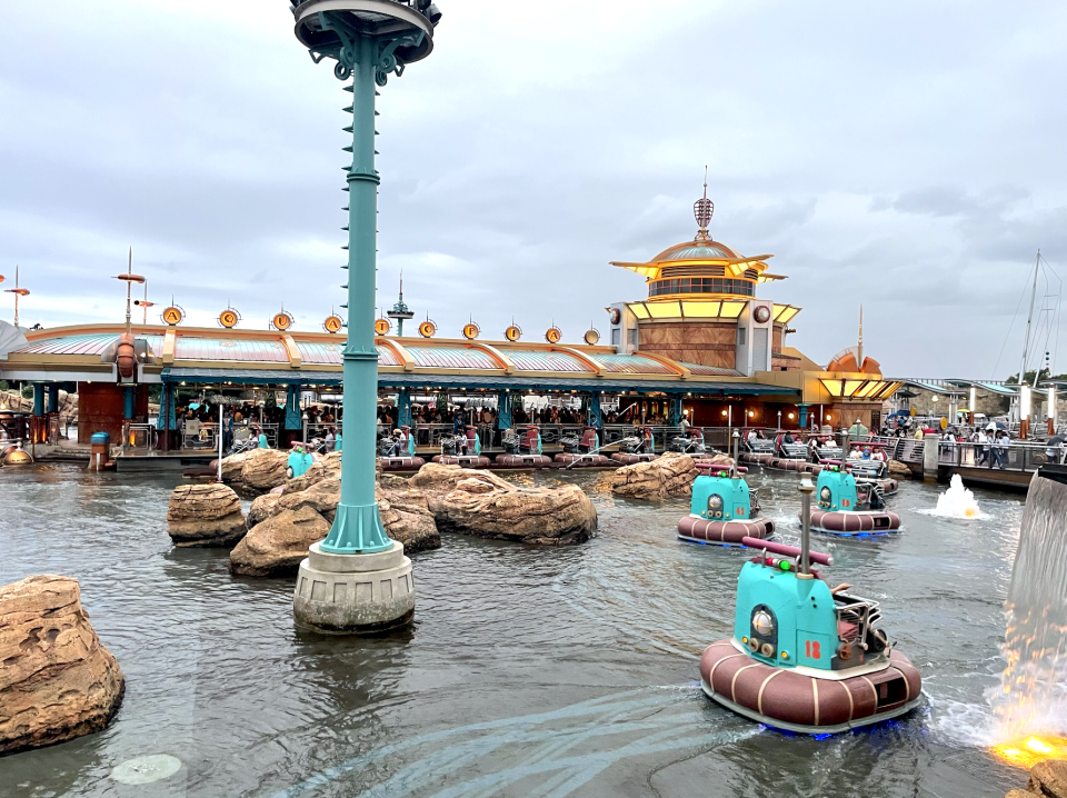 An amusement park water ride with boats designed like submarines navigating through rocky waters. In the background, a themed building appears with people inside