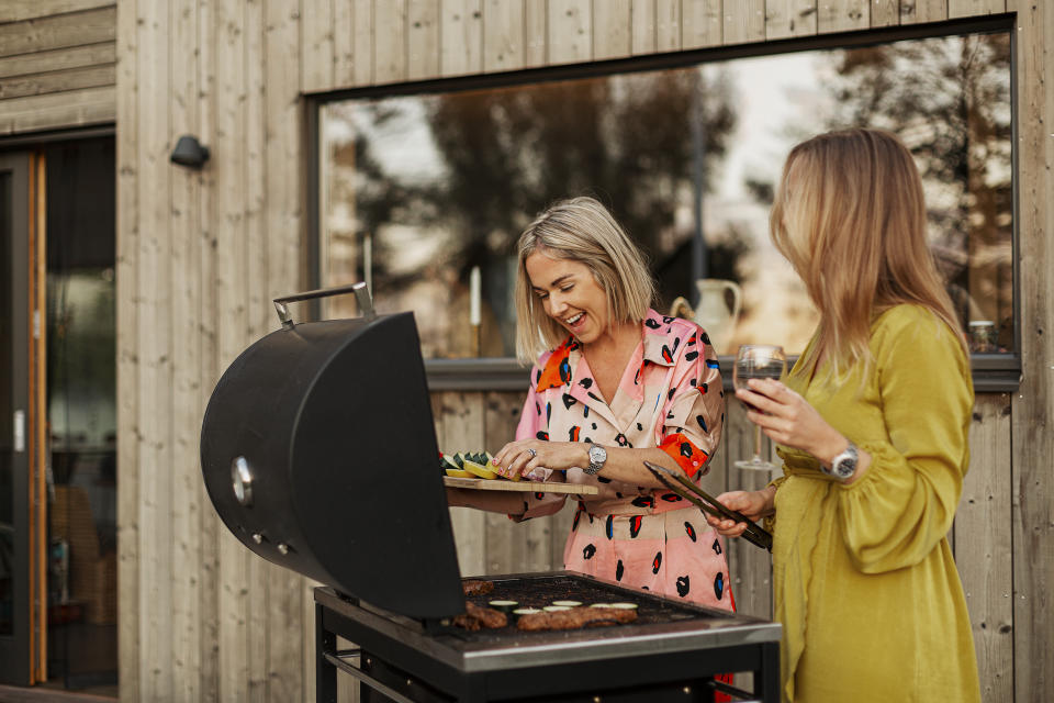 two women standing at black BBQ putting food on grill in front of wooden house