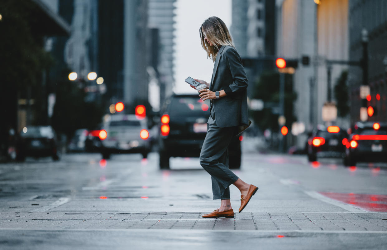 Businesswoman in formal attire walking on a city street while using her smartphone. Urban environment with traffic and buildings in the background.