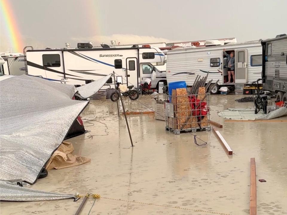 A couple of white RVs parked in a sea of mud with lawn chairs and loose pieces of wood everywhere. In the distance, two rainbows