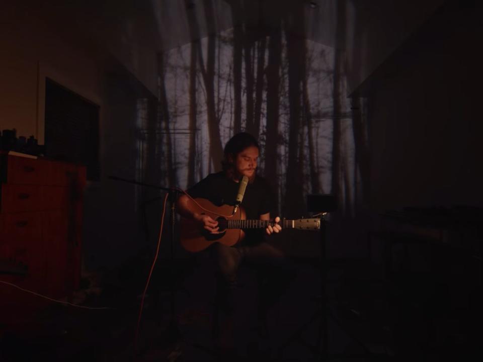 A man (Bo Burnham) playing an acoustic guitar while sitting in front of a projection of a forest background.