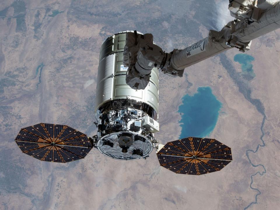 cygnus spacecraft silver cylinder with two orange and black circular solar arrays below a long robotic arm high above brown earth with lakes