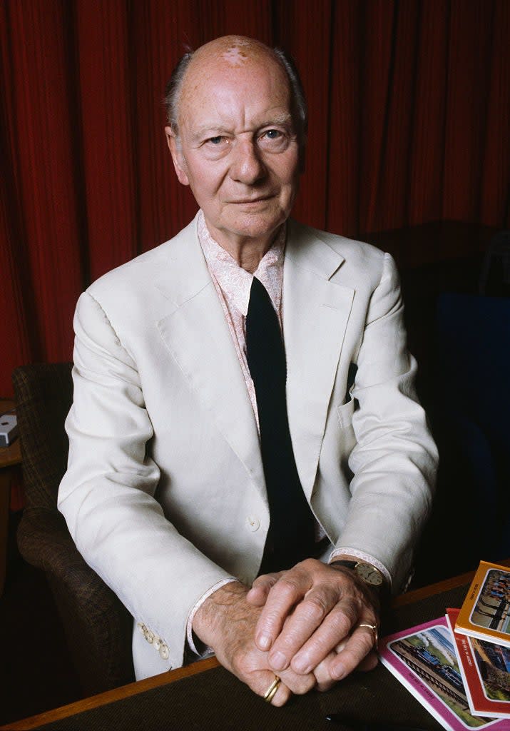 An older man is wearing a white suit jacket and black tie, with his hands clasped in front of him on a table with various colored cards