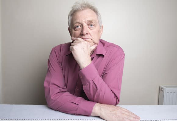 Older man sitting at a table with a ponderous expression as his chin rests on his hand.