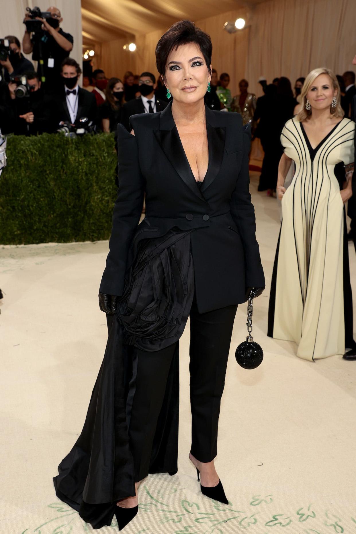 Kris Jenner attends the 2021 Met Gala wearing a black suit with a floral applique and plunging neckline. She accessorized with emerald earrings and a cannonball-shaped clutch.