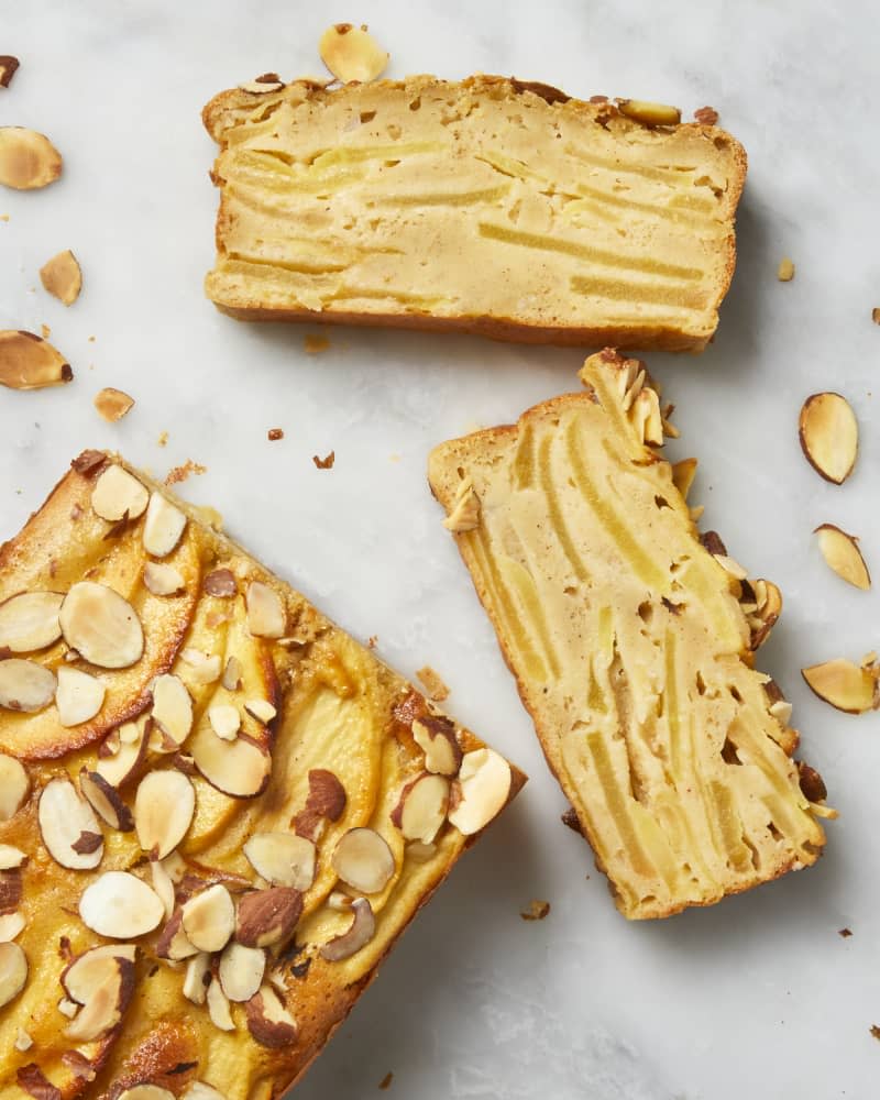 Overhead view of two slices and half of an uncut apple cake on a white surface with almonds scattered on the surface.