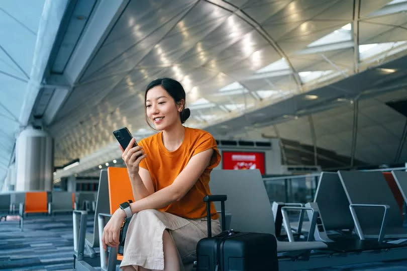 Young Asian woman with suitcase using smartphone while waiting for her flight at airport terminal.